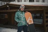 A woman in winter clothing stands confidently with her snowboard in front of a wooden cabin in a snowy setting.