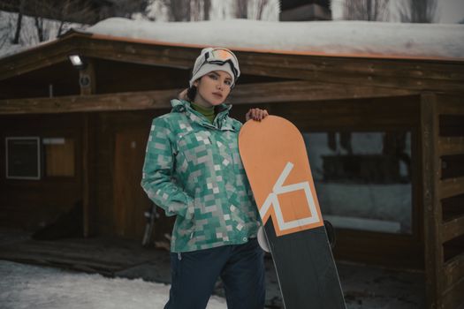 A woman in winter clothing stands confidently with her snowboard in front of a wooden cabin in a snowy setting.