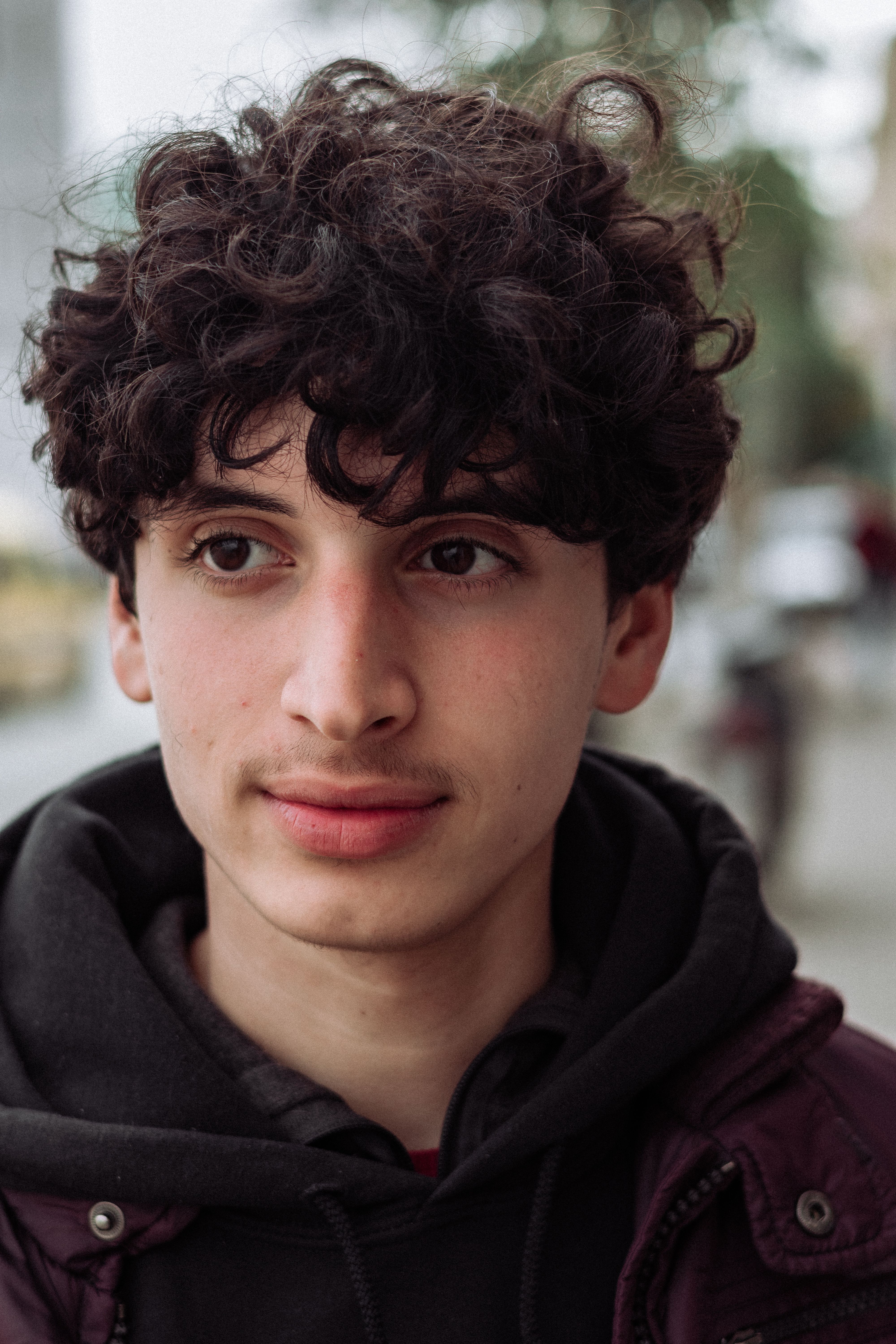 Portrait of a young man with curly hair wearing a jacket and hoodie outdoors.