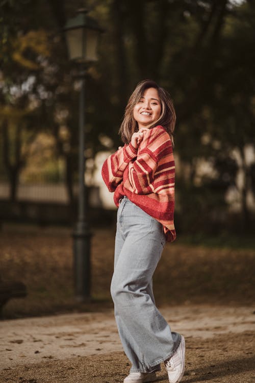 Young Woman in a Sweater and Jeans Standing in a Park and Smiling 