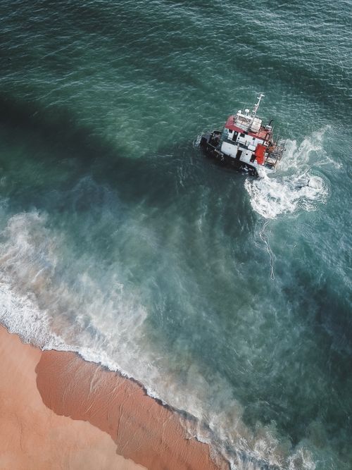 Free Drone shot of a sunken ship off the coast of Chukai, Terengganu, Malaysia showcasing ocean waves and sandy shore. Stock Photo