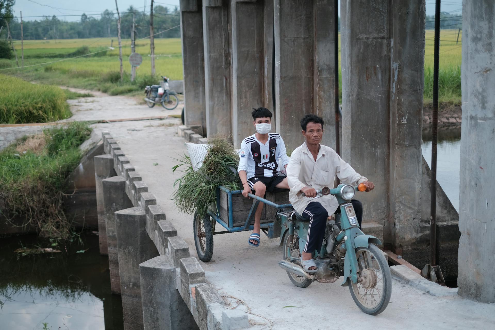 Men on Motorbike with Trailer on Footbridge in Village