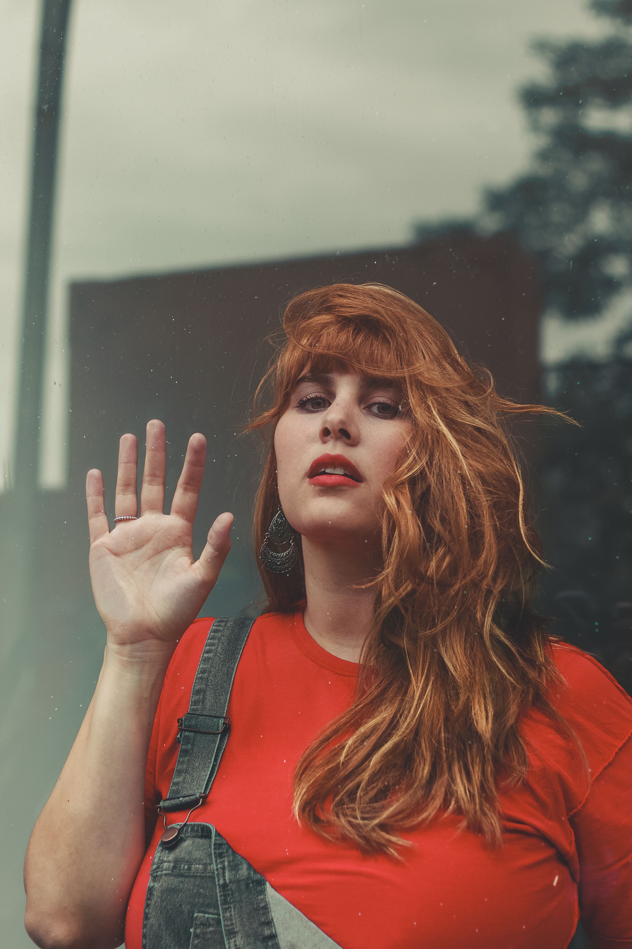 A young woman with red hair waves in a casual outdoor setting, wearing denim overalls and a red shirt.