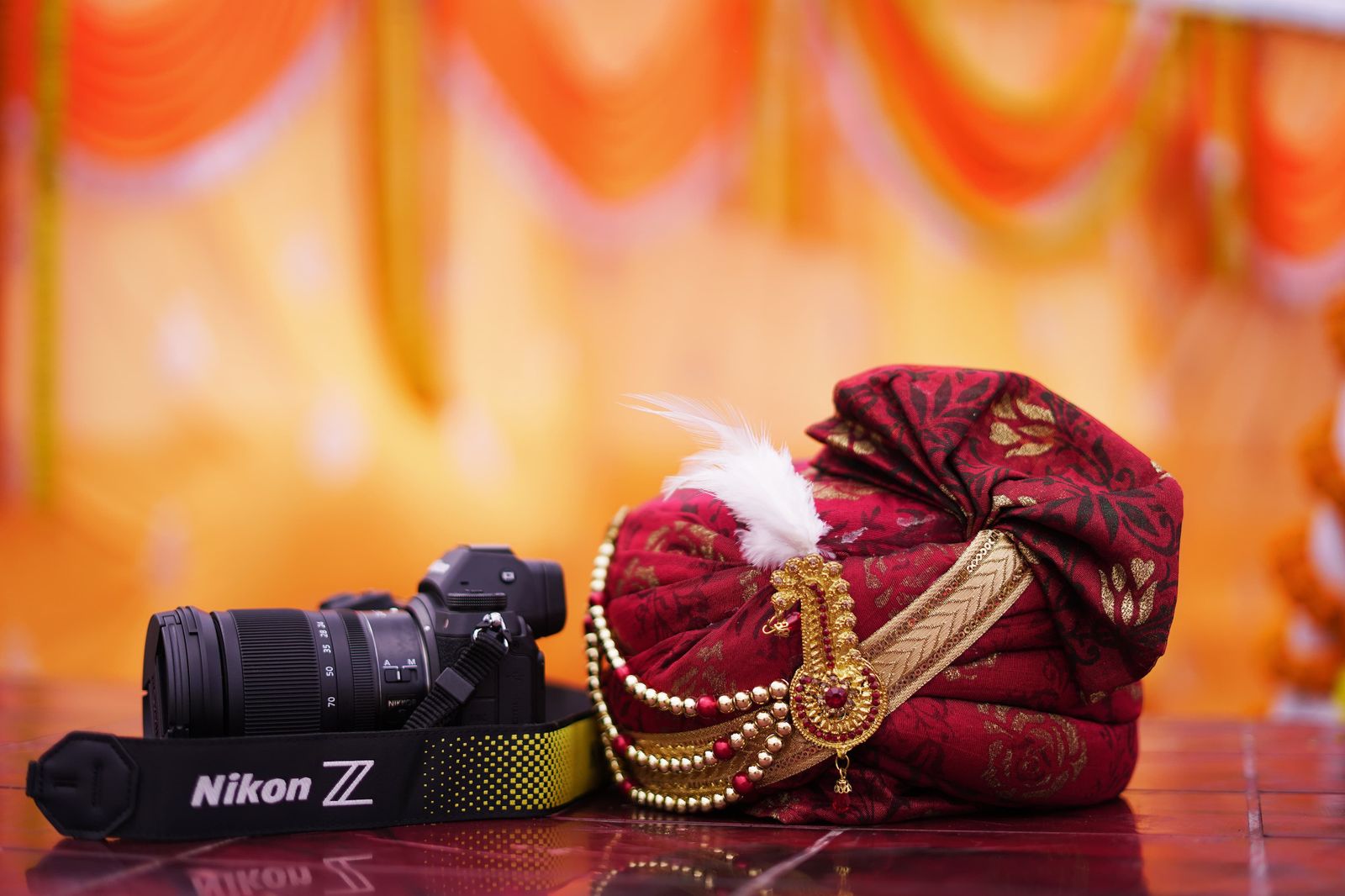 A maroon and gold brocade pagdi with a white kalgi and a gold sarpech, lying beside a camera on a polished floor, with marigold drapery thrown far out of focus behind it.