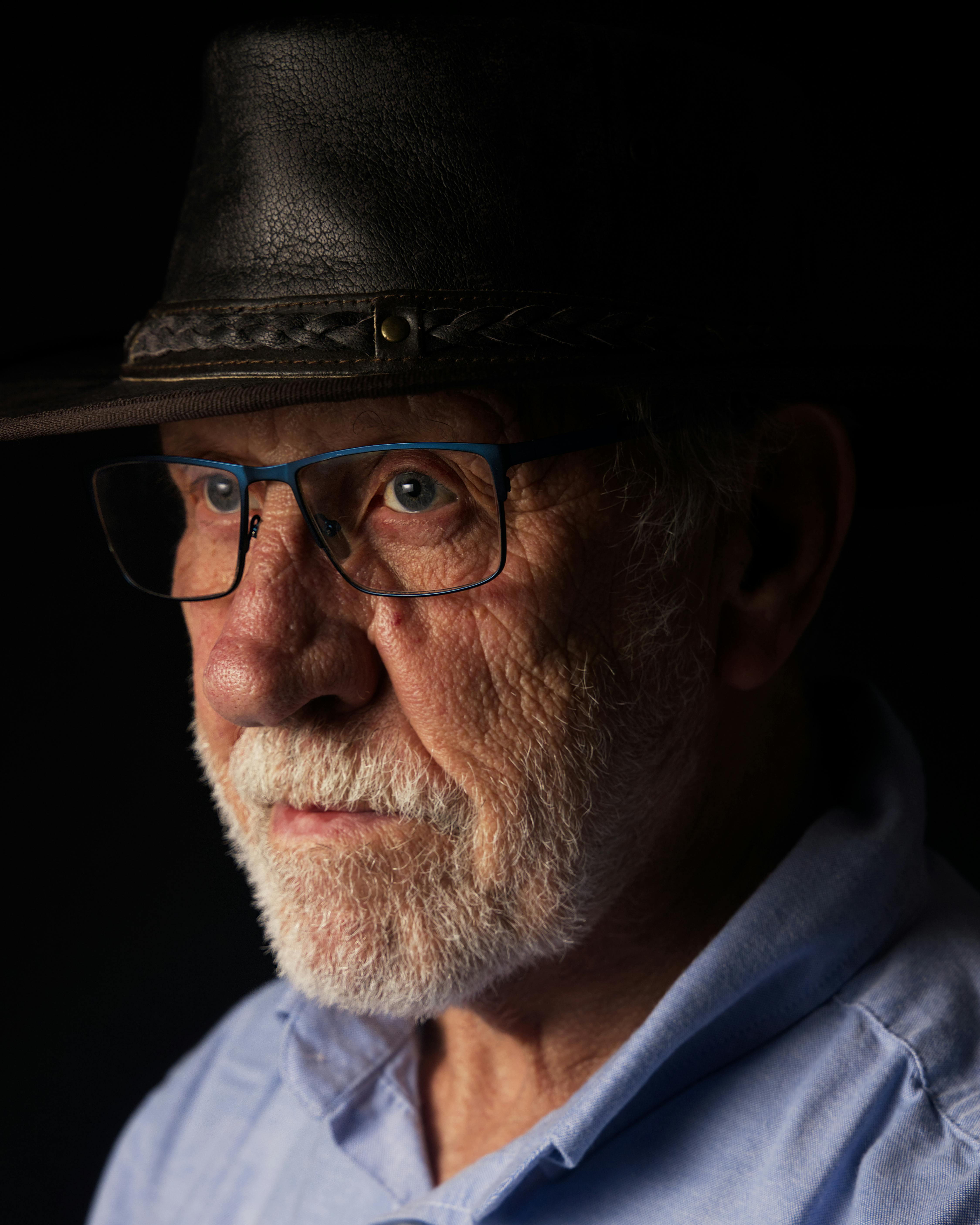 A detailed close-up portrait of an elderly man wearing a leather hat and eyeglasses against a black background.