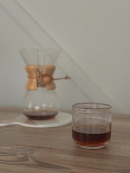 A glass of filter coffee on a wooden table with an elegant glass coffee maker in the background.