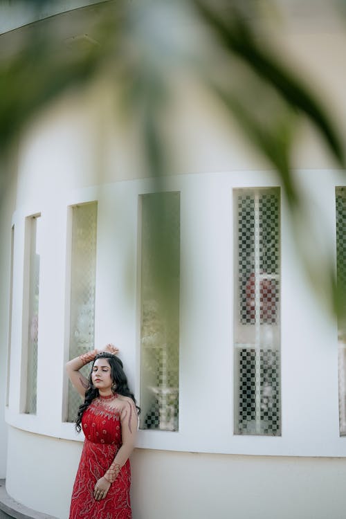 Young Woman in an Elegant Red Dress Standing with Closed Eyes in Front of the House Windows