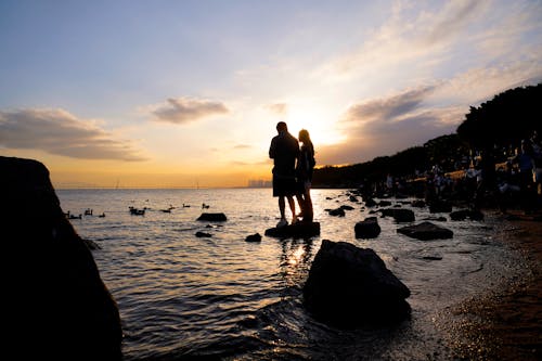 Silhouettes of a Man and Woman Standing on a Rock on the Shore at Sunset