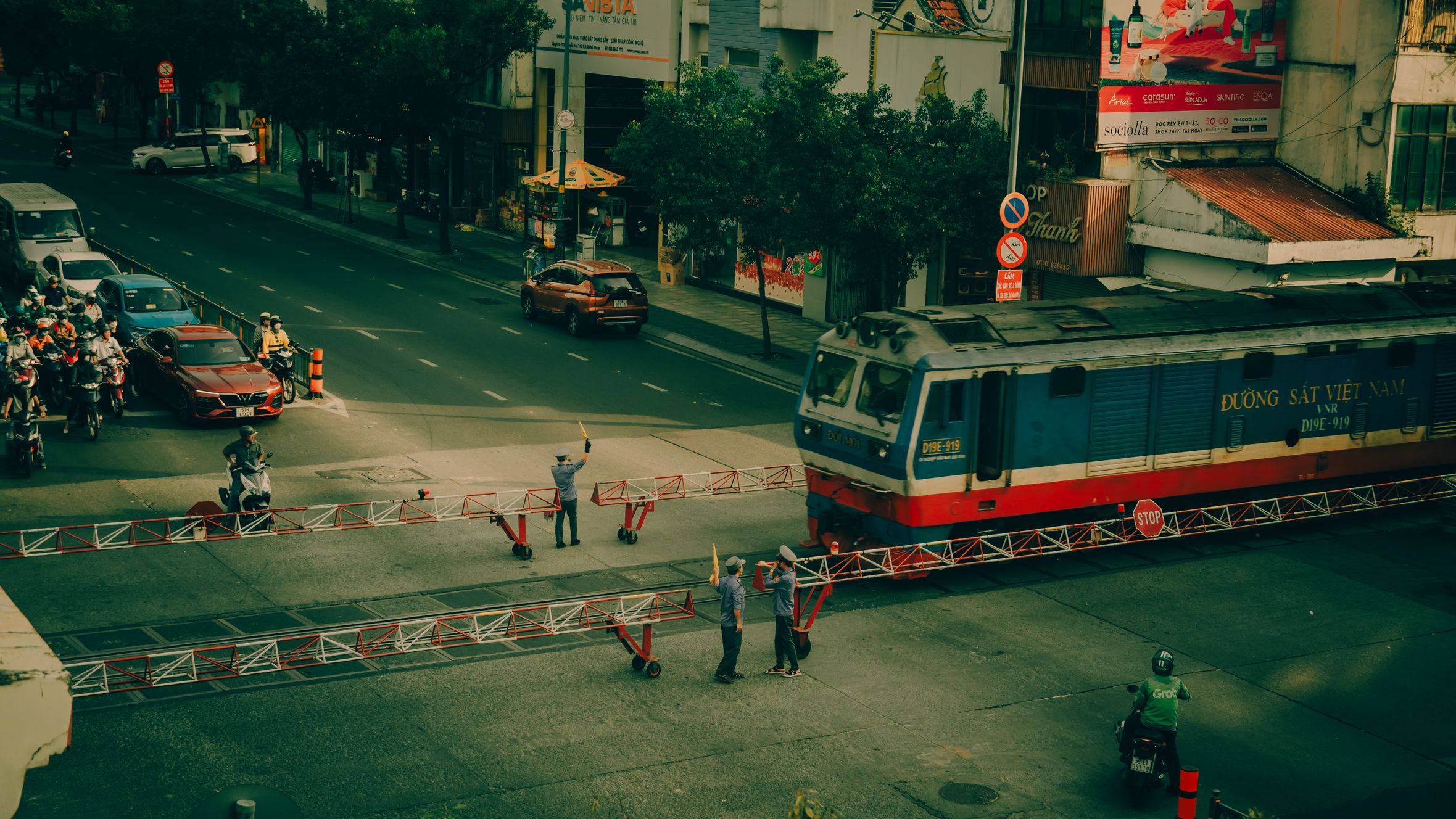 A Vietnamese city street showing a train crossing, with visible signage and city traffic.