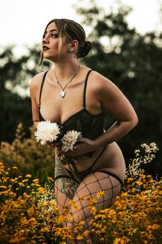 A woman in black lingerie posing with flowers in a scenic outdoor garden setting.
