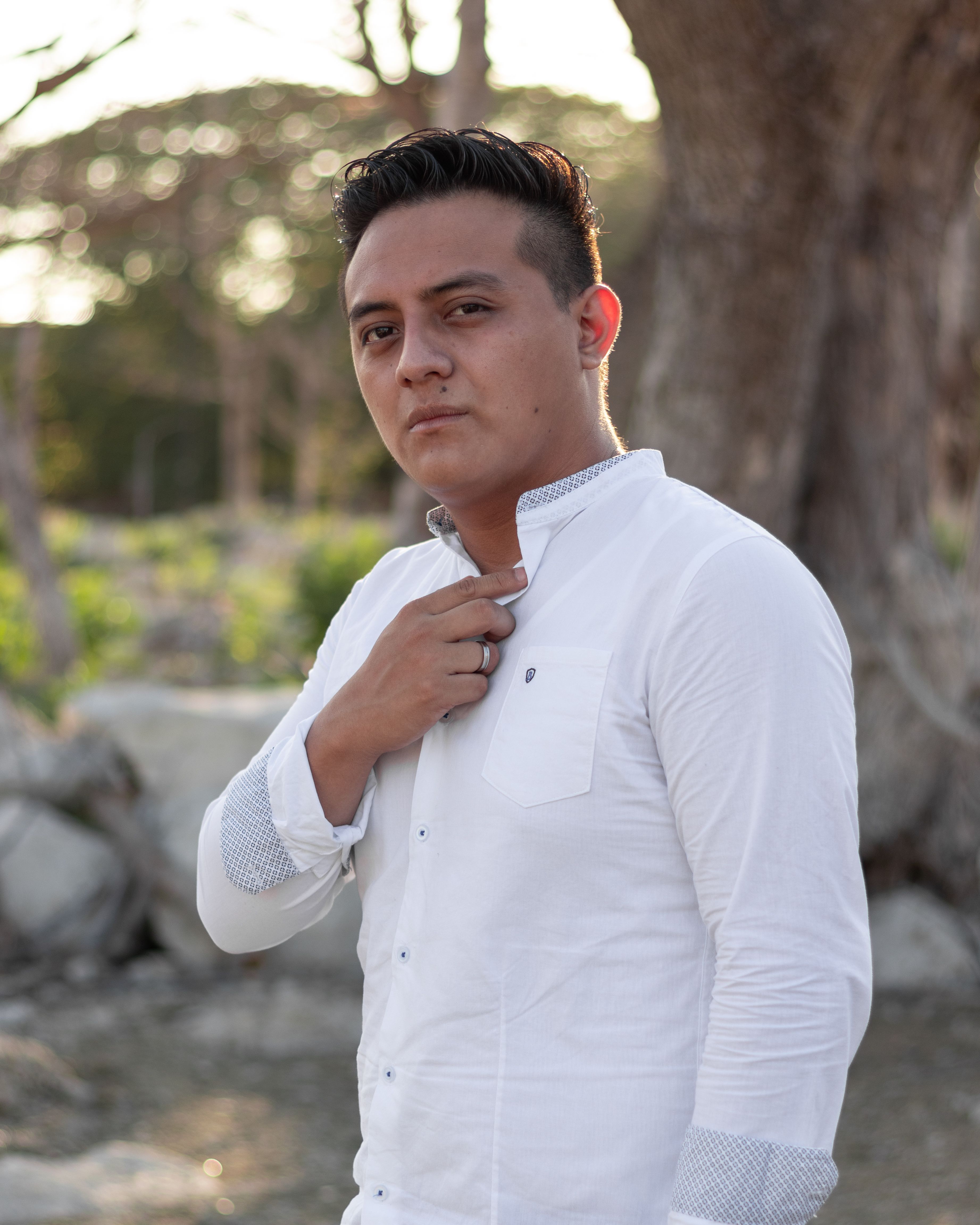 A confident man standing outdoors in a white shirt, captured in soft natural light.