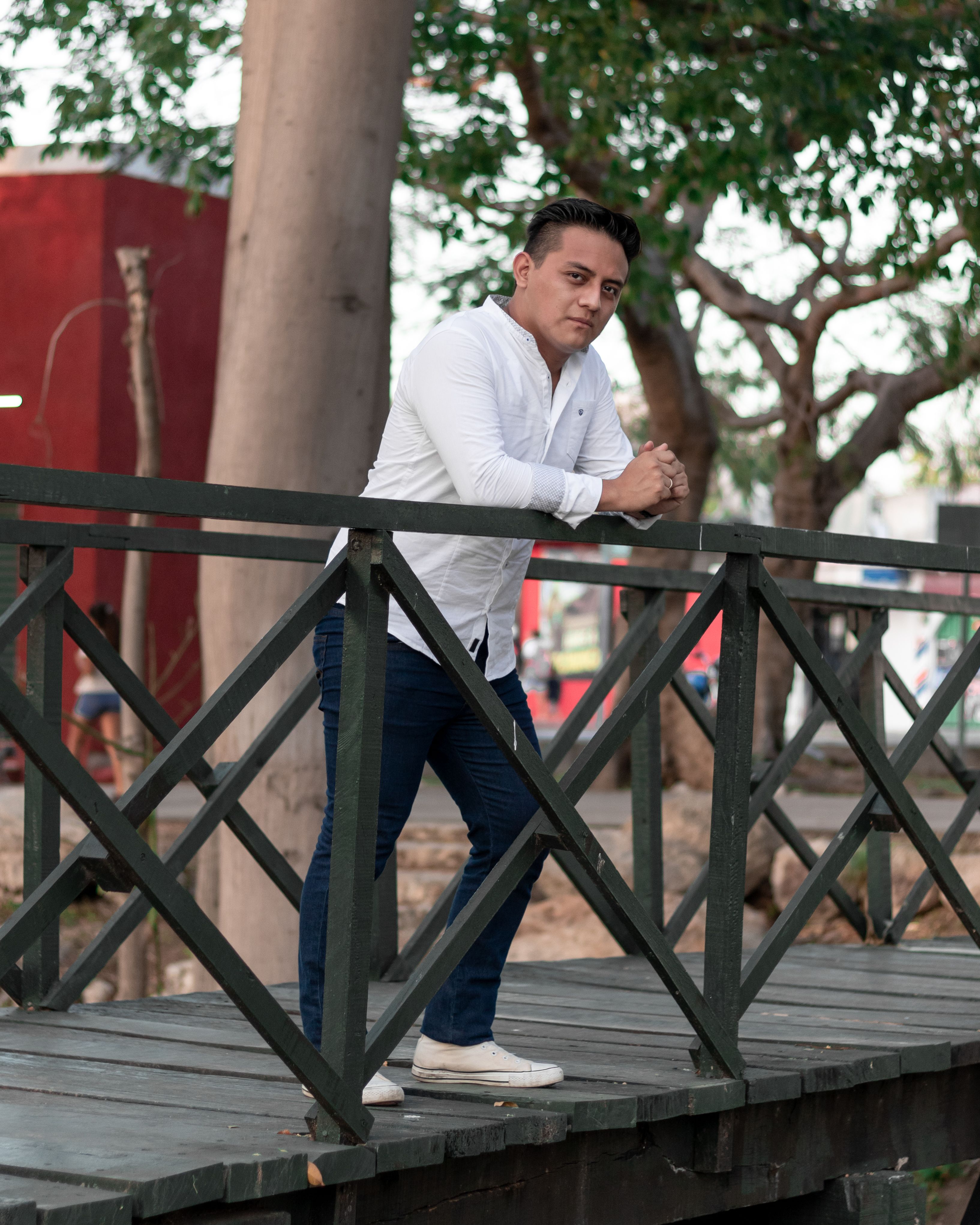 Portrait of a man in a white shirt leaning on a wooden footbridge outdoors.