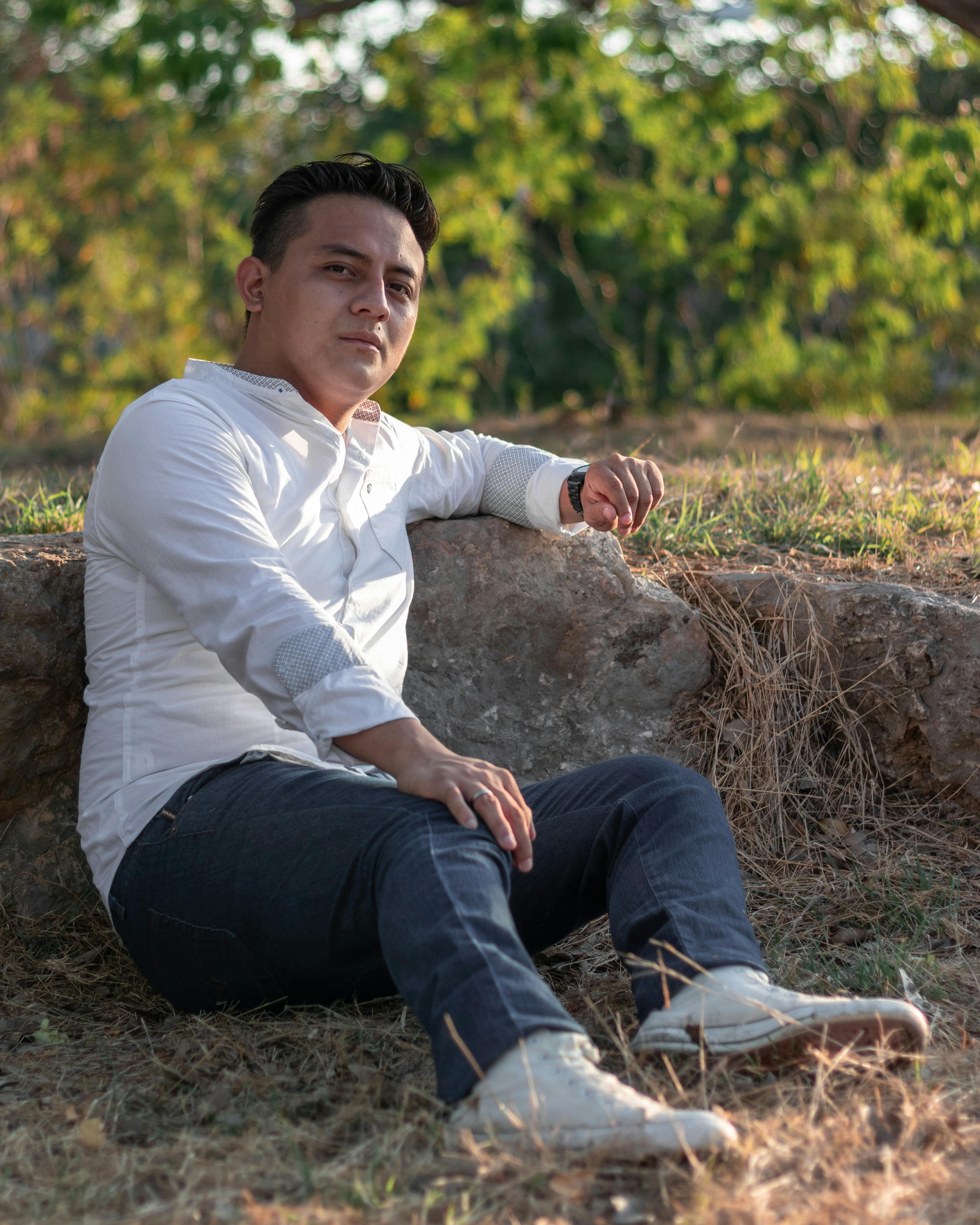 A man in a white shirt and jeans sits on a rock outdoors, enjoying a sunny day.
