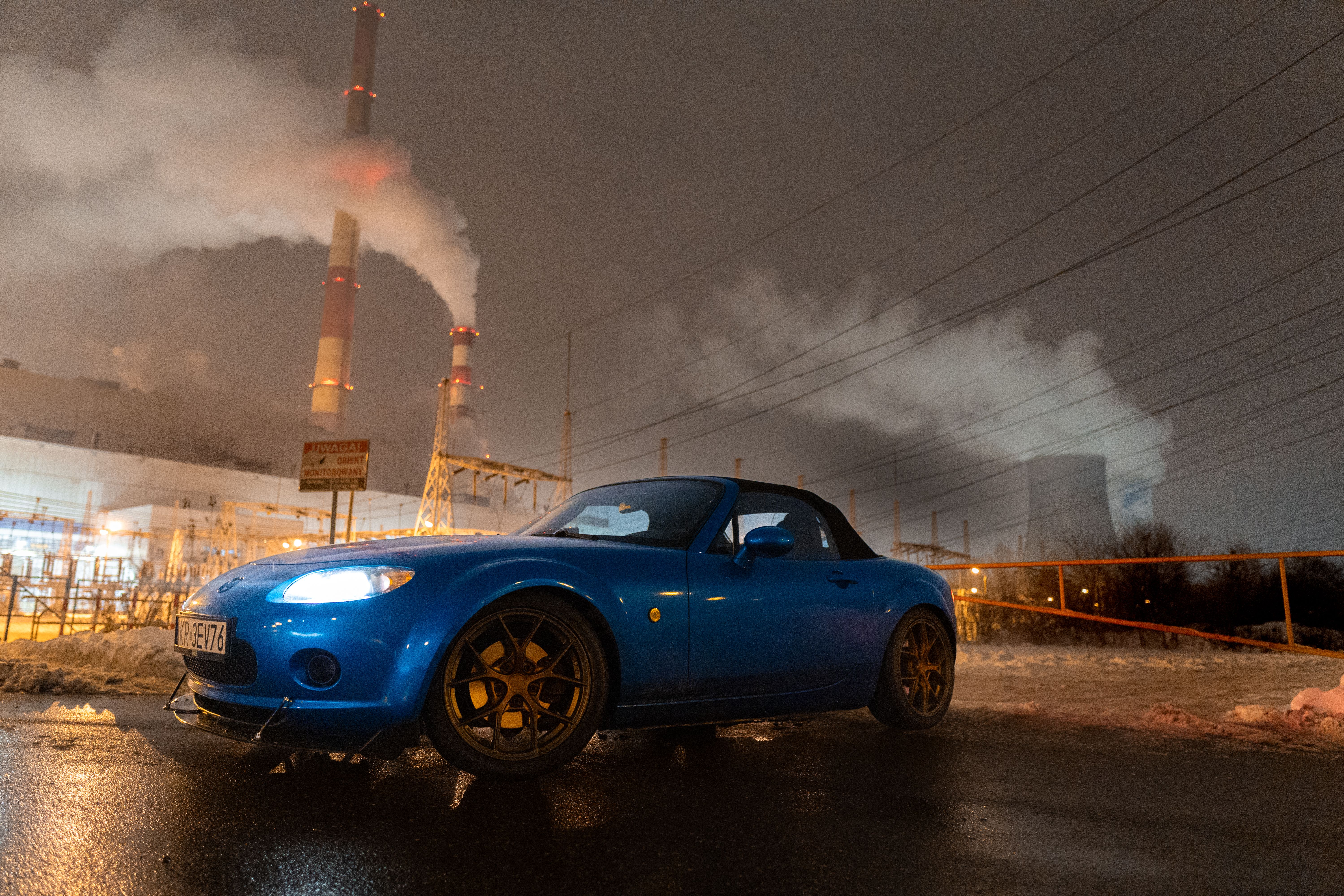 Blue sports car parked near a smoking industrial plant in Kraków at night.