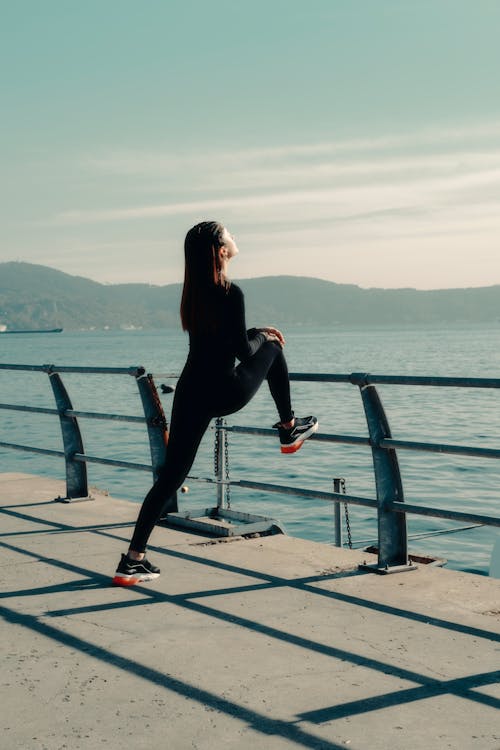 Woman in Sportswear Stretching Her Legs against the Railing along the Shore 