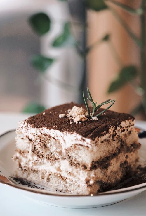 Free Close-up of a delicious tiramisu cake slice with creamy layers, topped with cocoa and herb garnish on a plate. Stock Photo