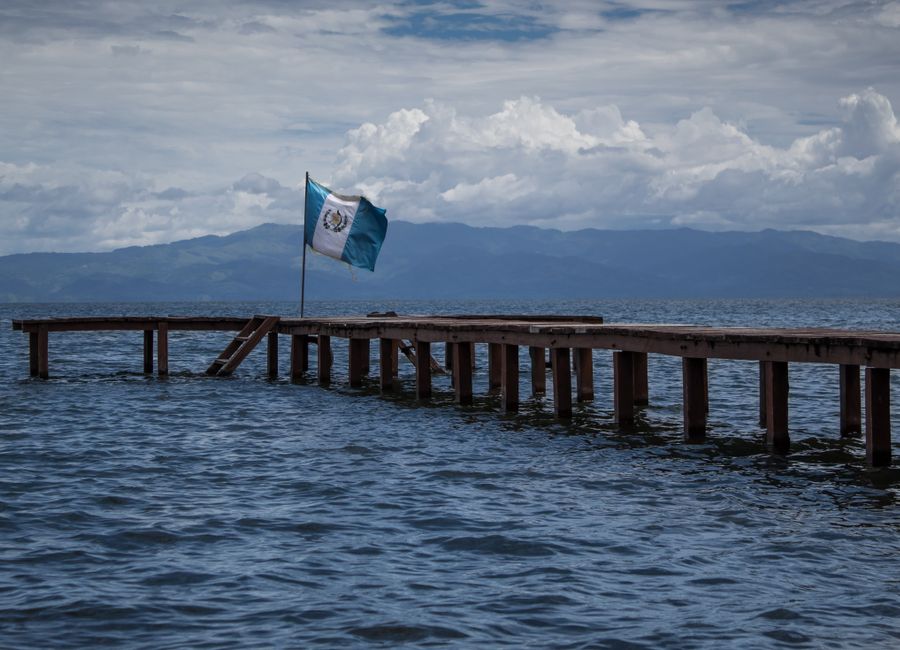 Wooden pier extending into a lake with the Guatemalan flag waving, set against a backdrop of mountains and a cloudy sky.