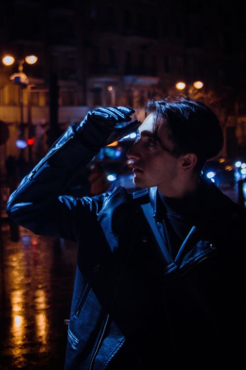 Free A fashionable man wearing a black jacket and gloves poses confidently in a vibrant nighttime street scene. Stock Photo