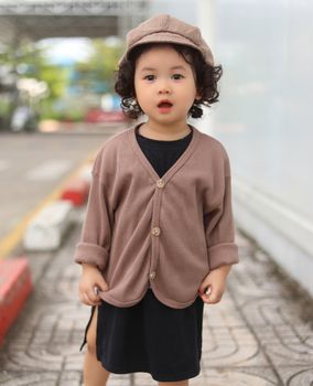 Charming young girl in a brown cardigan and hat posing in an urban setting.