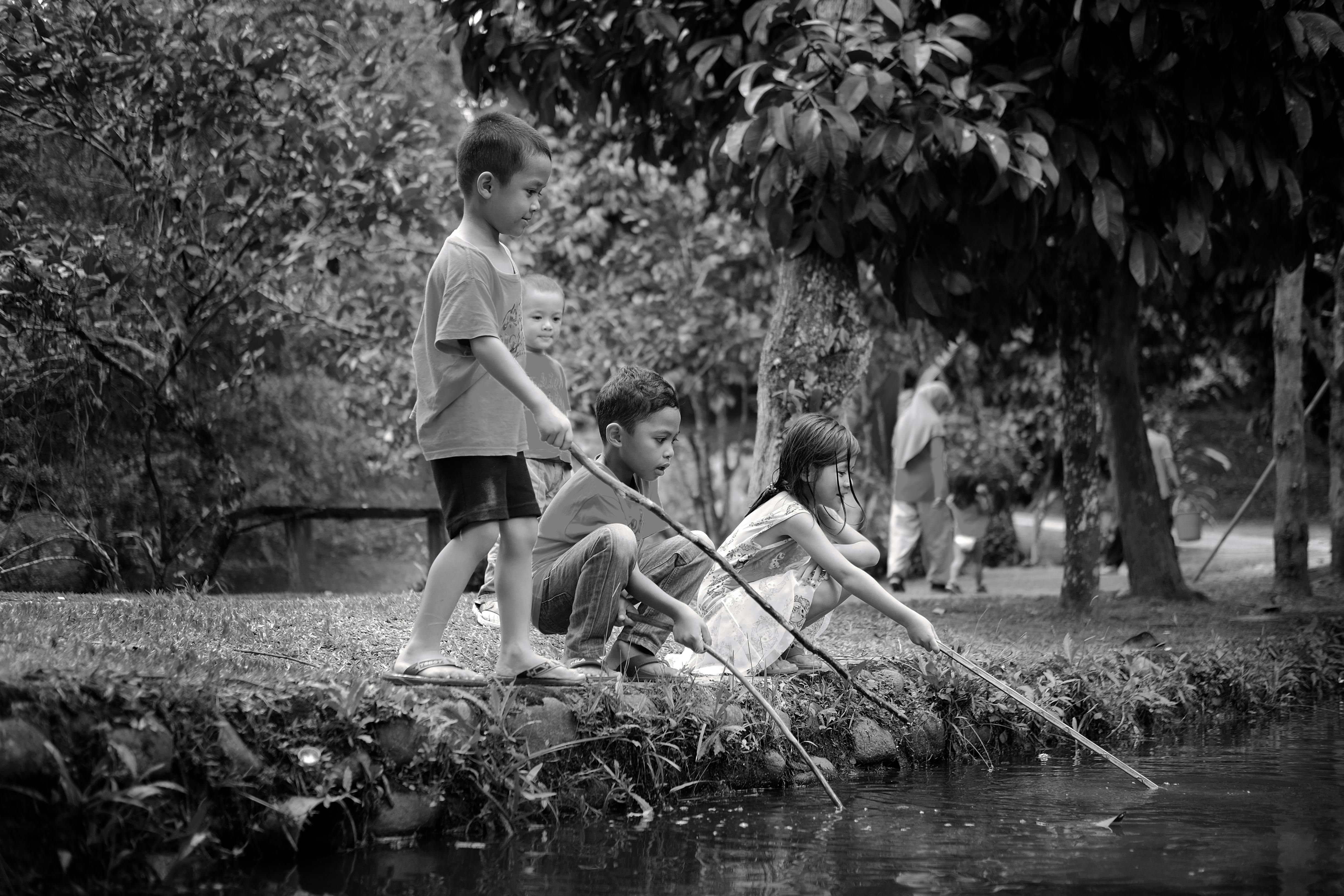 Black and white photo of children playing with sticks by a pond in a park.
