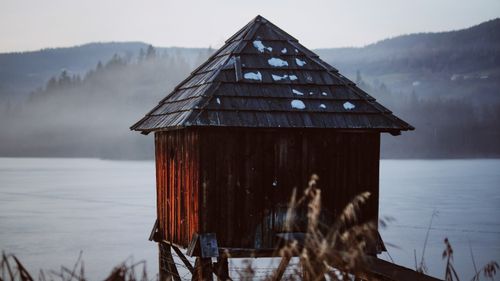 Free A rustic wooden hut stands by a misty lake with foggy hills in the background. Stock Photo