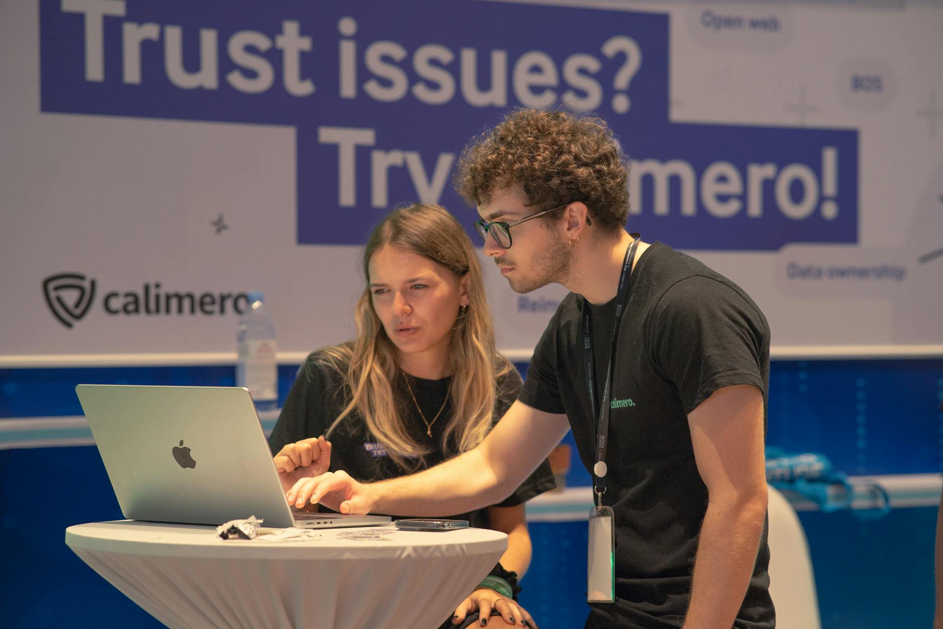 Two individuals collaborating on a laptop at a tech conference booth promoting Calimero.