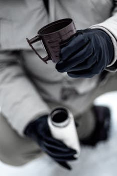Close-up of a person with gloves holding a cup and flask in a snowy setting.