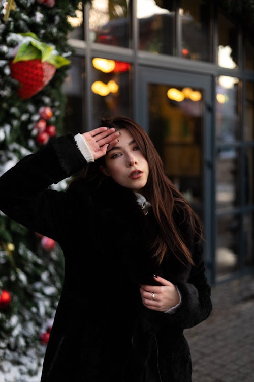 Young Model in a Black Fur Coat with Her Hand Placed on Her Forehead Standing on the Sidewalk by the Christmas Tree