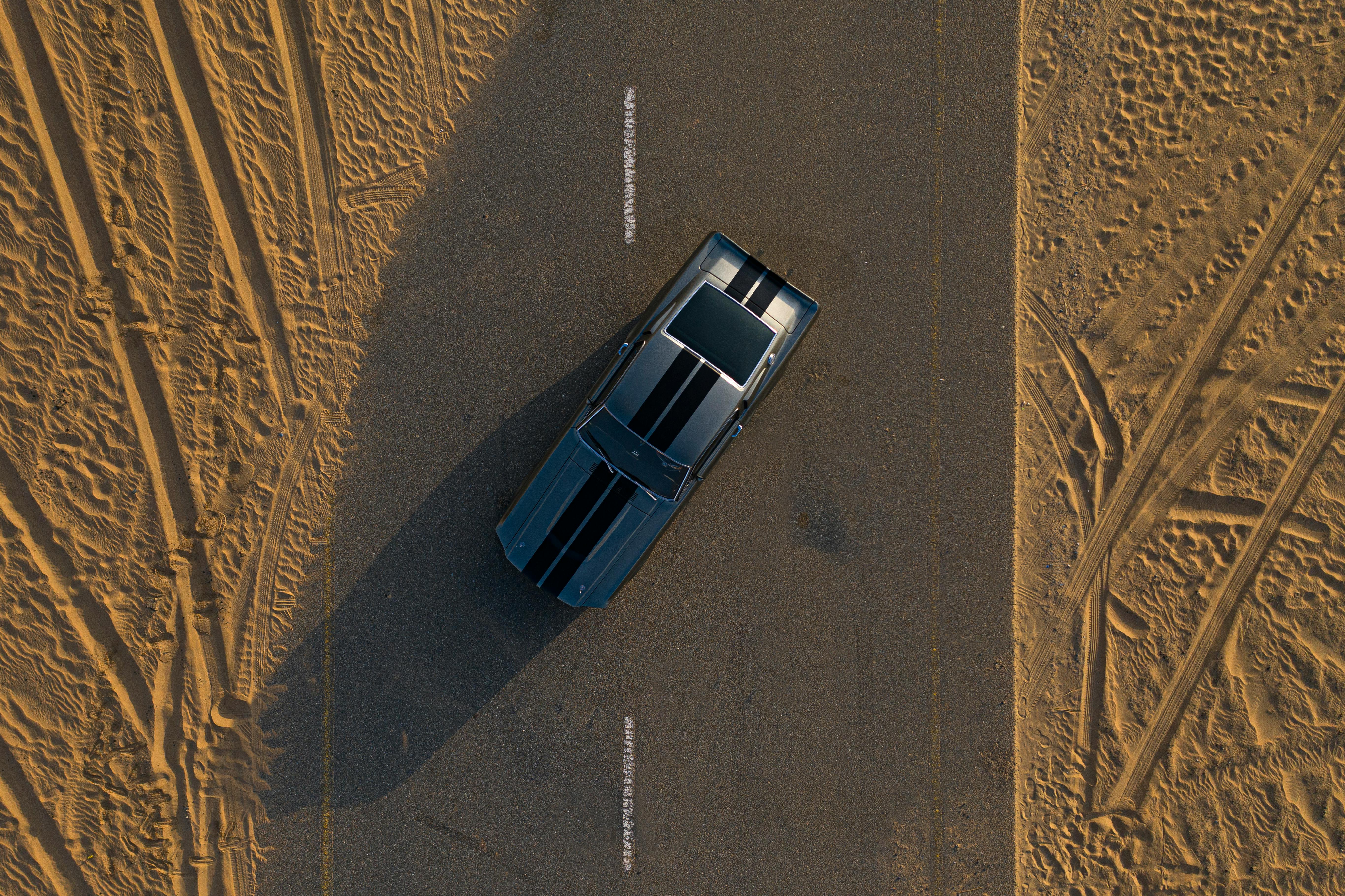 Drone shot of a black muscle car on a deserted road with sand tracks.
