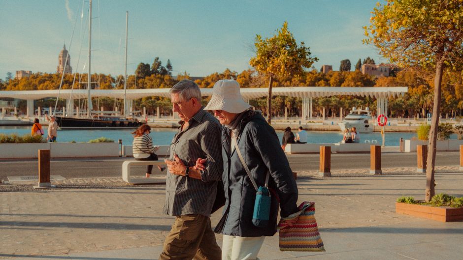 Pareja de mayores camina abrazada a lo largo del soleado paseo en Málaga, España. Escenario urbano pintoresco.