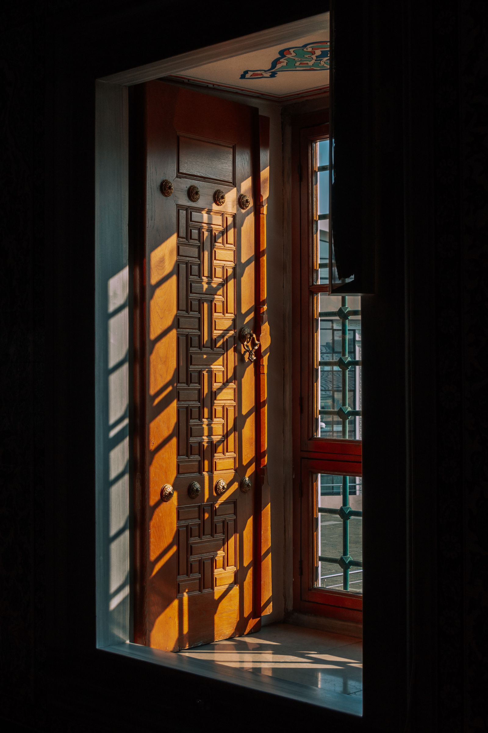 Warm sunlight filtering through an ornate wooden window shutter, casting geometric shadows indoors