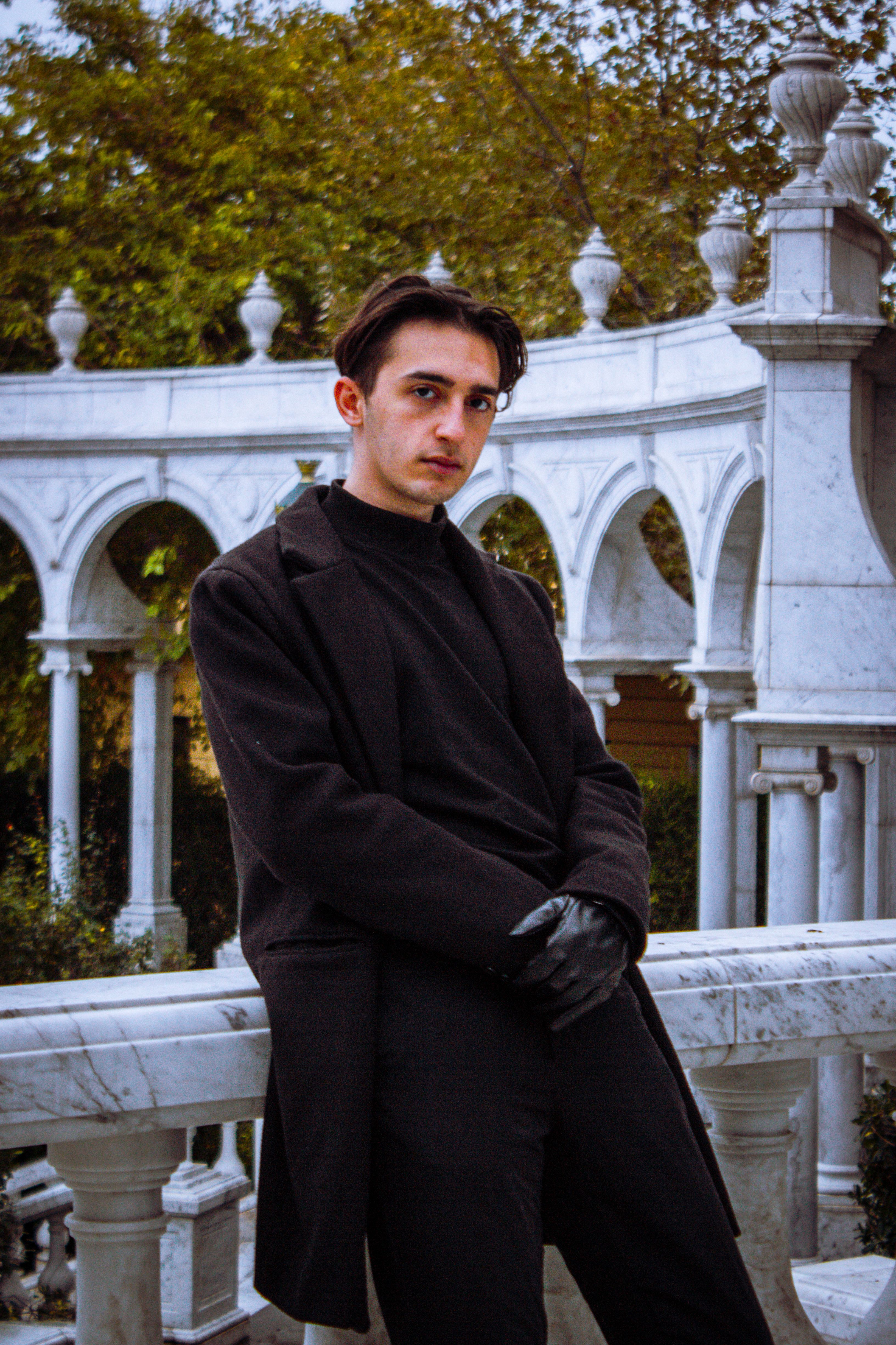 Stylish man in black coat leaning against marble balustrade in an outdoor classical setting.