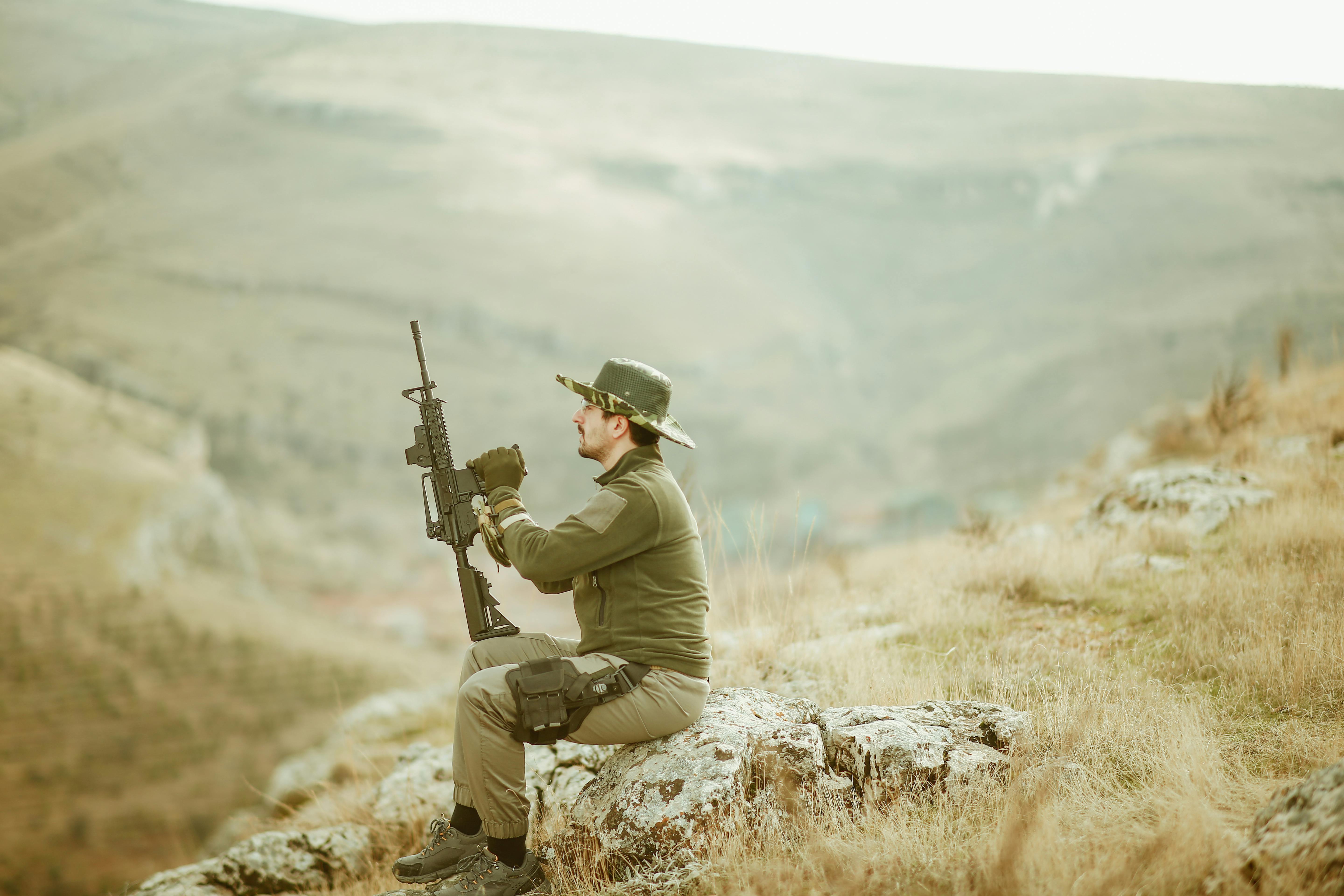 A man holding a rifle sits on a rock in the Malatya mountains, Türkiye, showcasing outdoor adventure.
