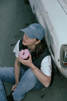 Young woman in casual attire sitting by a vintage car, holding a pink doughnut.