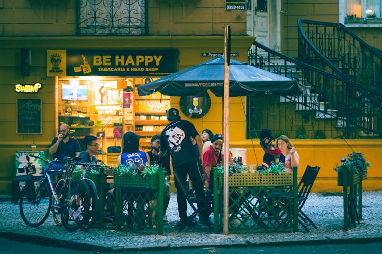 People Sitting Under An Umbrella In Front Of A Shop 