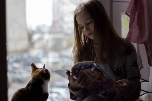 Girl in jumper with cat pattern reading magazine quietly while sitting on windowsill with cute curious cat attentively watching