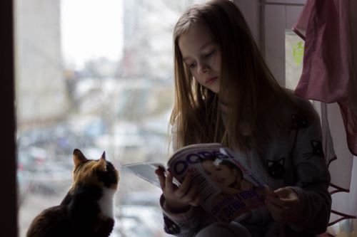 Free Girl in jumper with cat pattern reading magazine quietly while sitting on windowsill with cute curious cat attentively watching Stock Photo