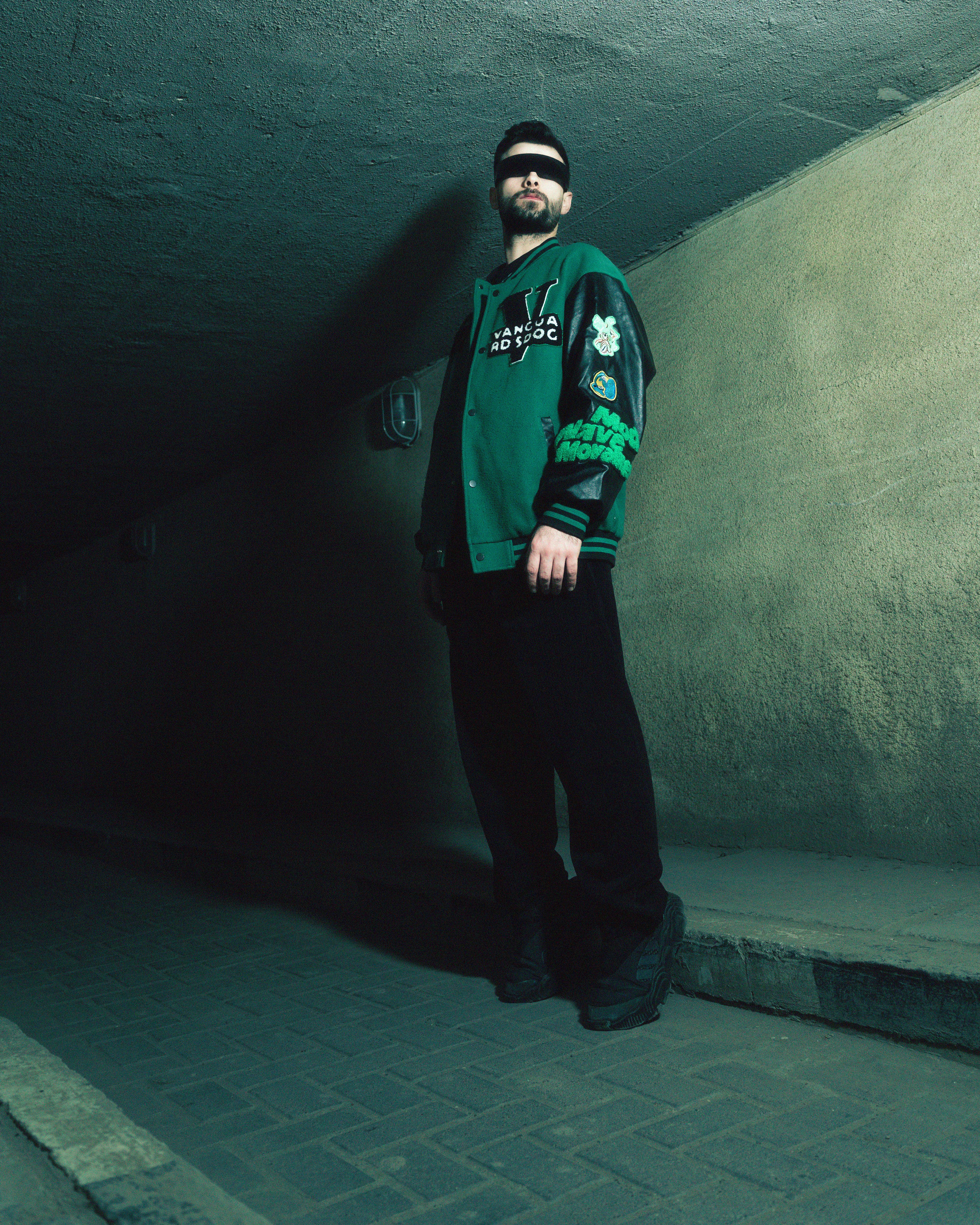 Portrait of a man in a blindfold and green jacket in an urban underpass, moody fashion vibe.