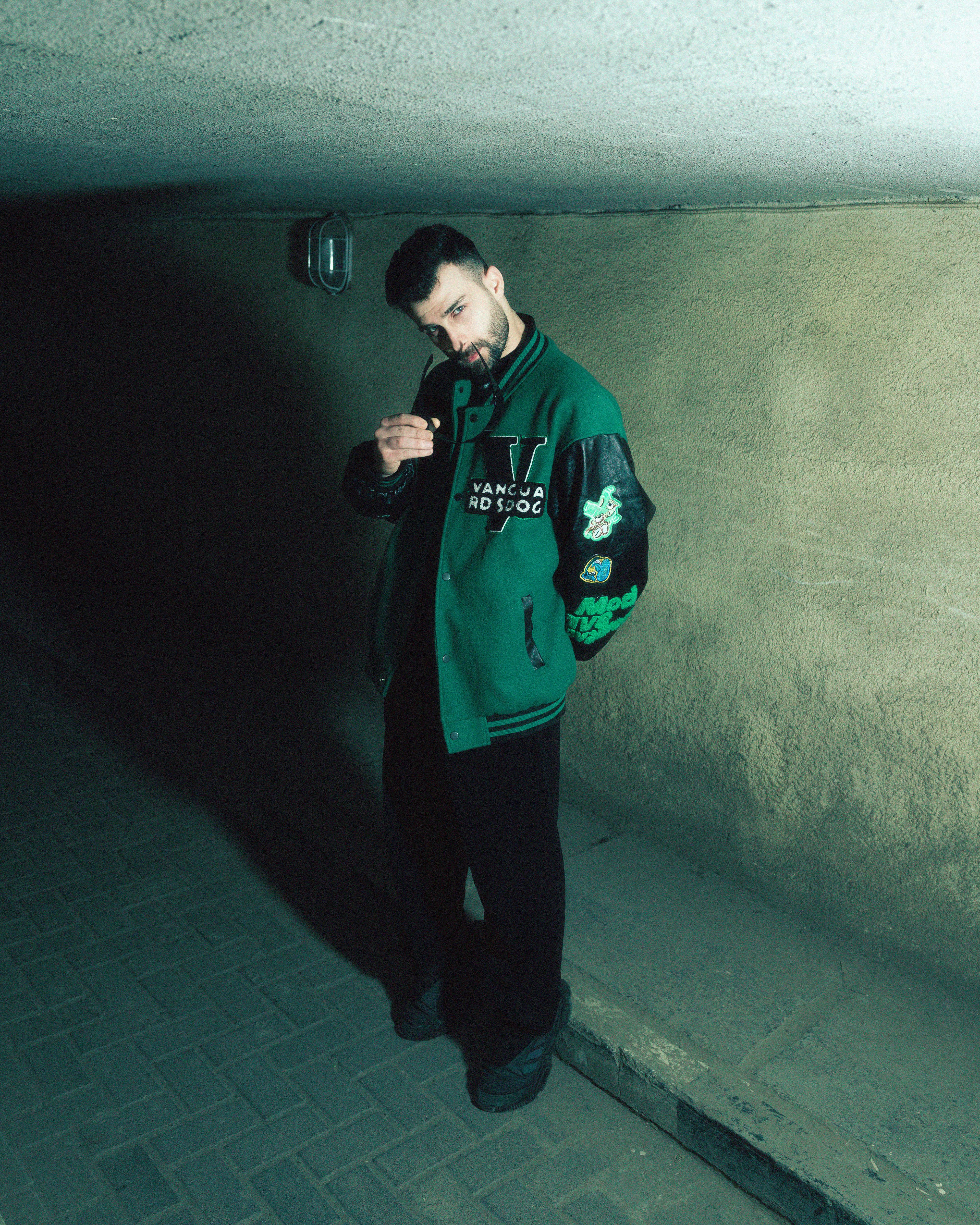 A man poses confidently in an underground tunnel wearing a stylish vintage varsity jacket.