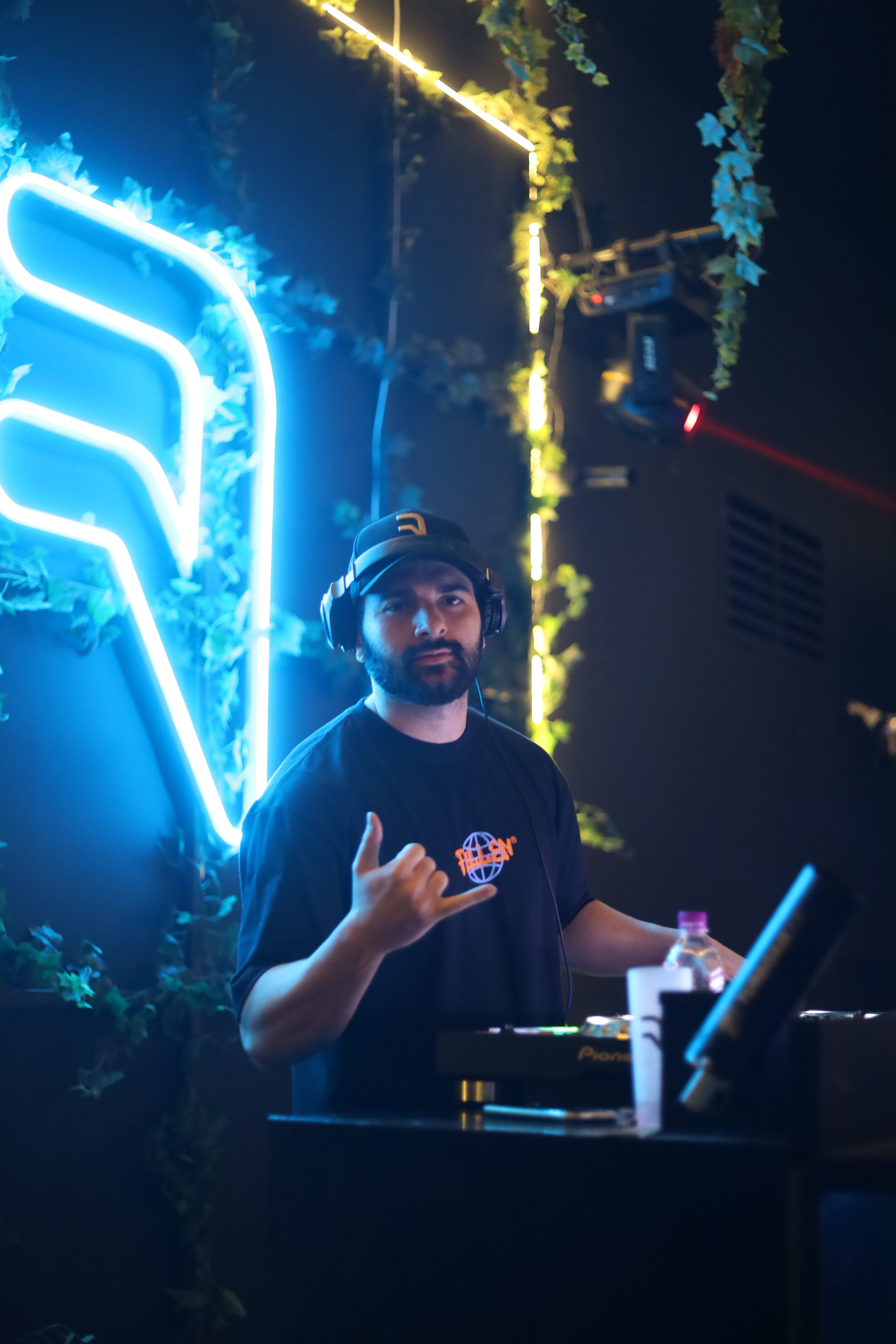 A DJ gestures with a shaka sign under neon lights in a vibrant nightclub setting, creating a lively atmosphere.