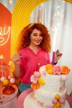 A joyful woman with curly red hair smiles beside a decorated birthday cake.