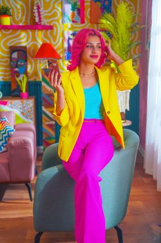 A confident woman with pink hair poses stylishly in a vibrant, colorful room.
