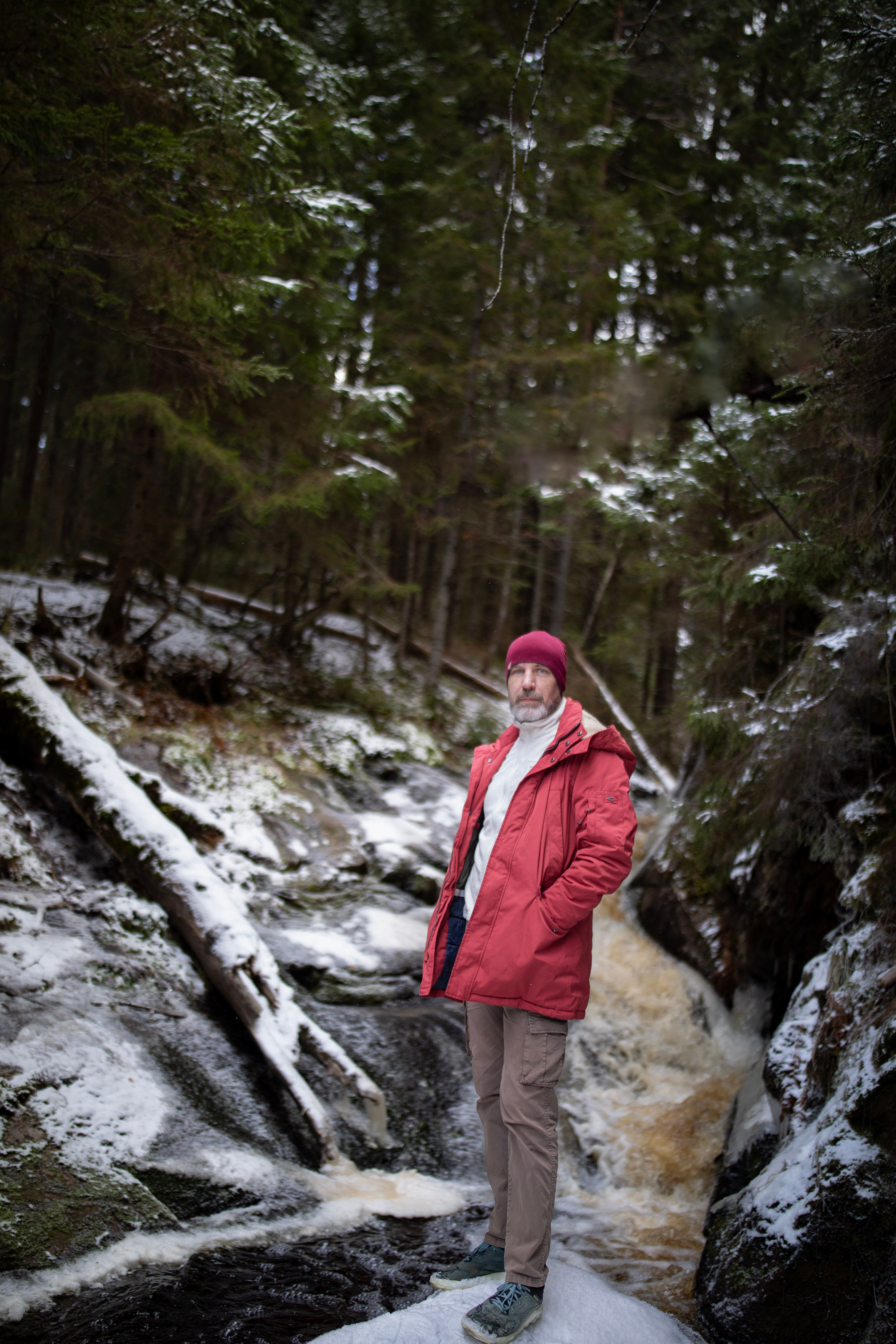 A man in winter attire stands confidently near a scenic snowy forest creek.