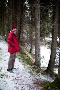 A man in a red jacket stands on a snowy forest path, surrounded by trees.