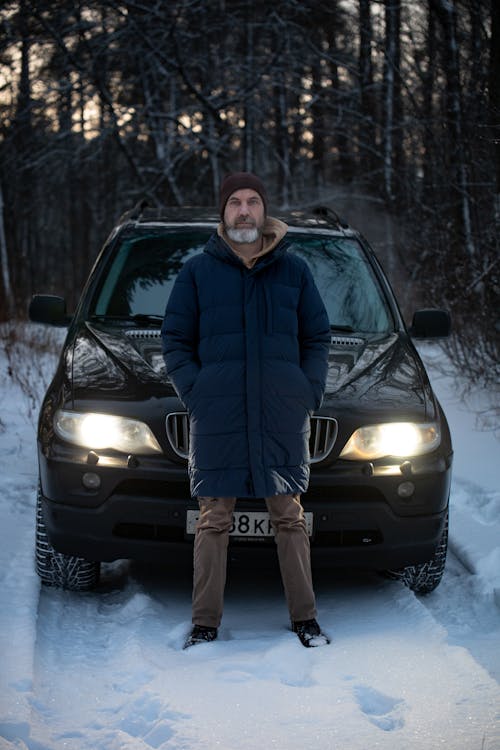 Man Standing against a Car with Switched on Headlights, in Winter Landscape