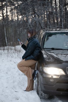 A woman leans on a car, using a smartphone in a snowy winter forest setting.