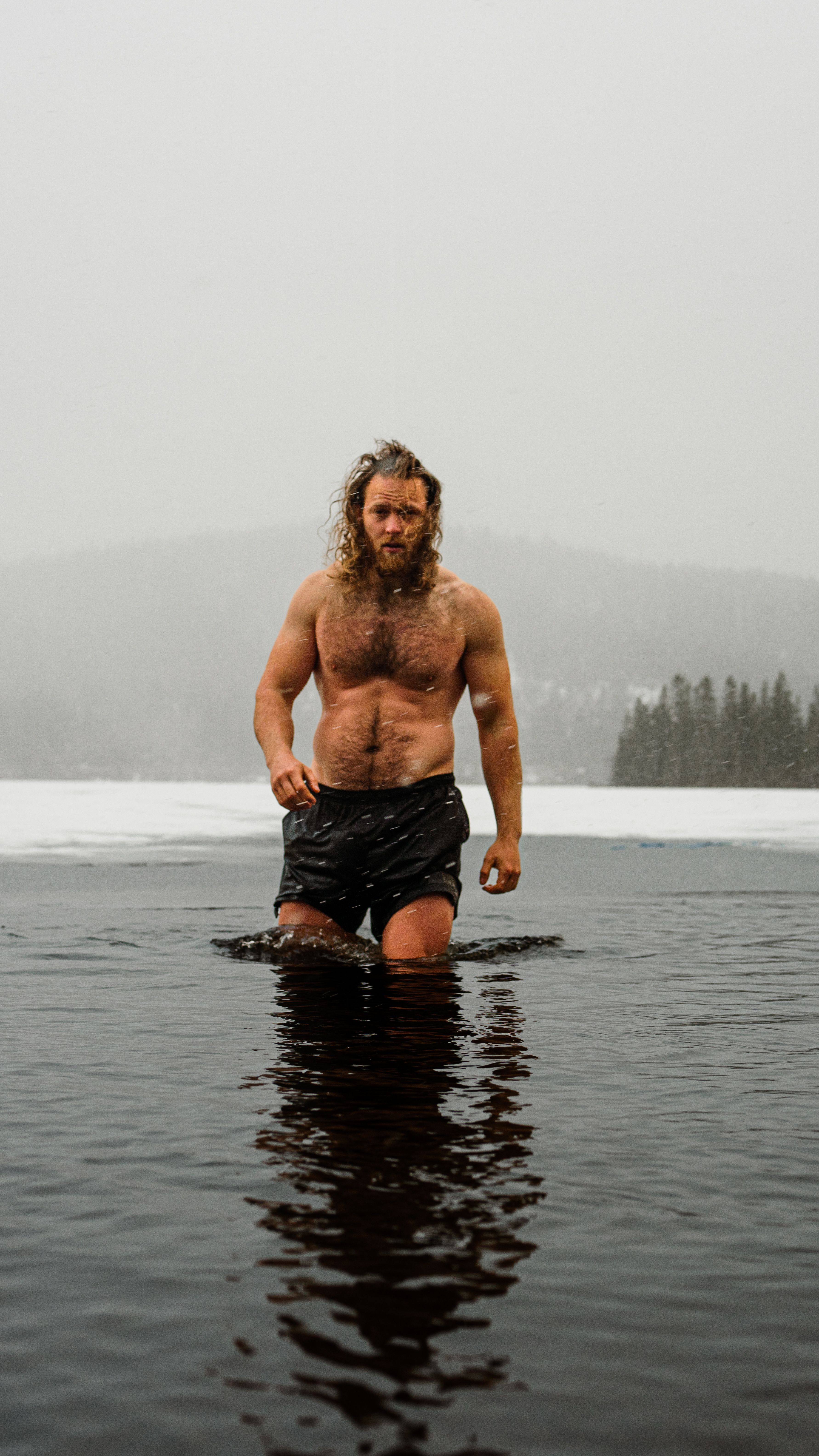 A topless bearded man braves icy lake water in a winter wilderness, embodying adventure.