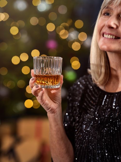 Free A woman in a sparkling dress enjoys a glass of whiskey with festive bokeh lights in the background. Stock Photo