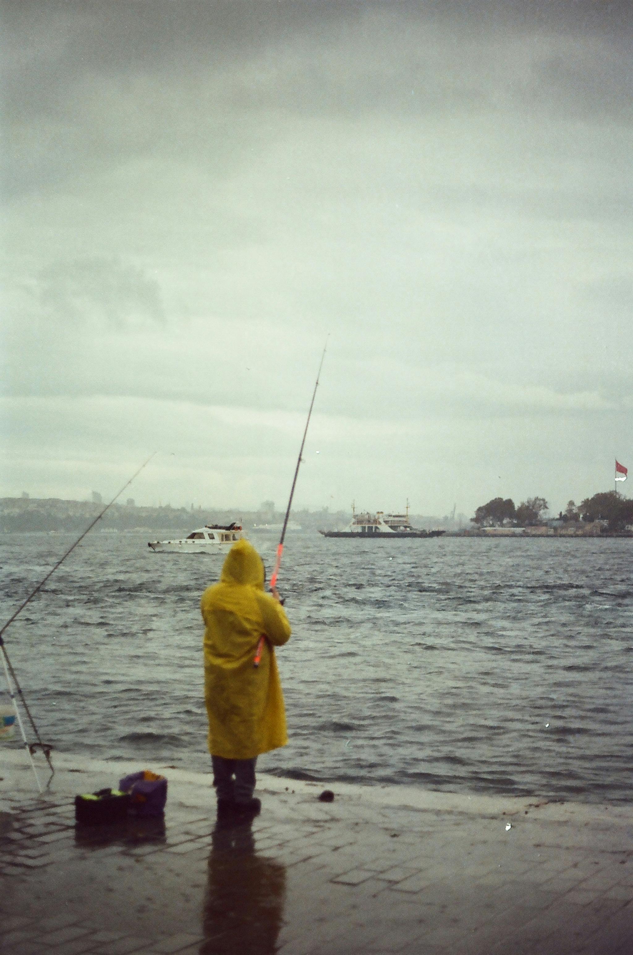Fisherman in a yellow raincoat casts a line into a stormy sea with gray skies.
