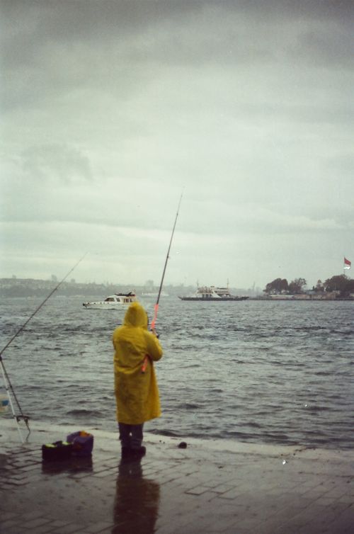 Free Fisherman in a yellow raincoat casts a line into a stormy sea with gray skies. Stock Photo